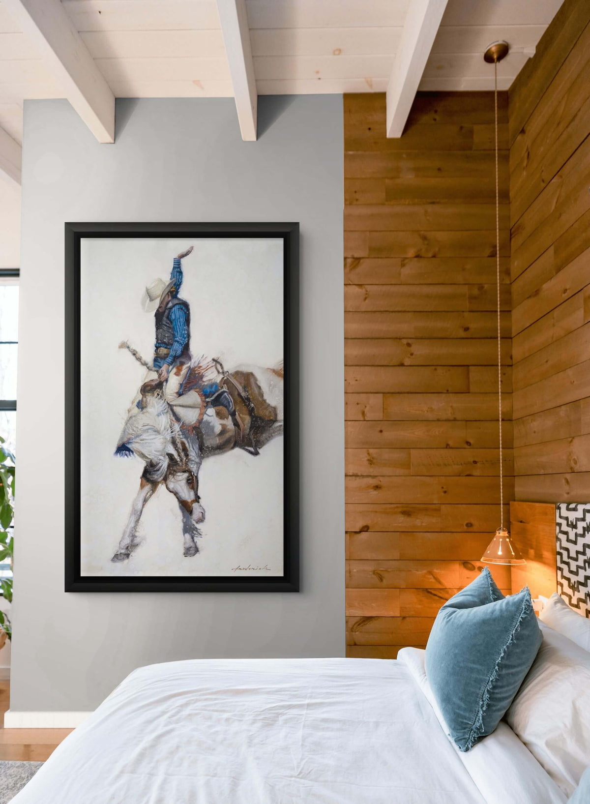 A framed art print depicting a cowboy riding a bucking bull, displayed on a gray wall in a rustic bedroom setting with a wooden slat headboard, white bedding, and blue pillows.